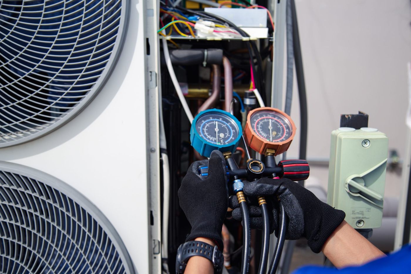 HVAC technician holding blue and orange pressure gauges while servicing air conditioning unit